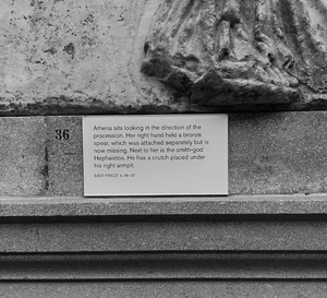 In the exhibit space, each section of the frieze has a placard, which describes the scene depicted on the section of frieze as well as the cardinal direction and number in the frieze series. The label for the east frieze V, 36- 37 reads as follows: "Athena sits looking in the direction of the procession. Her right hand held a bronze spear, which was attached separately but is not missing. Next to her is the smith-god Hephaistos. He has a crutch places under his right armpit."