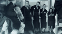 Photograph of multiple people standing in a room while a man in a hat plays the accordion at the 300th General Hospital in Naples, Italy.