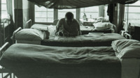 Photograph of a man with a cast looking at playing cards on a bed in the 300th General Hospital in Naples, Italy.