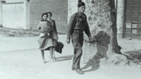 Photograph of a soldier followed by two women, one of whom is holding a baby.
